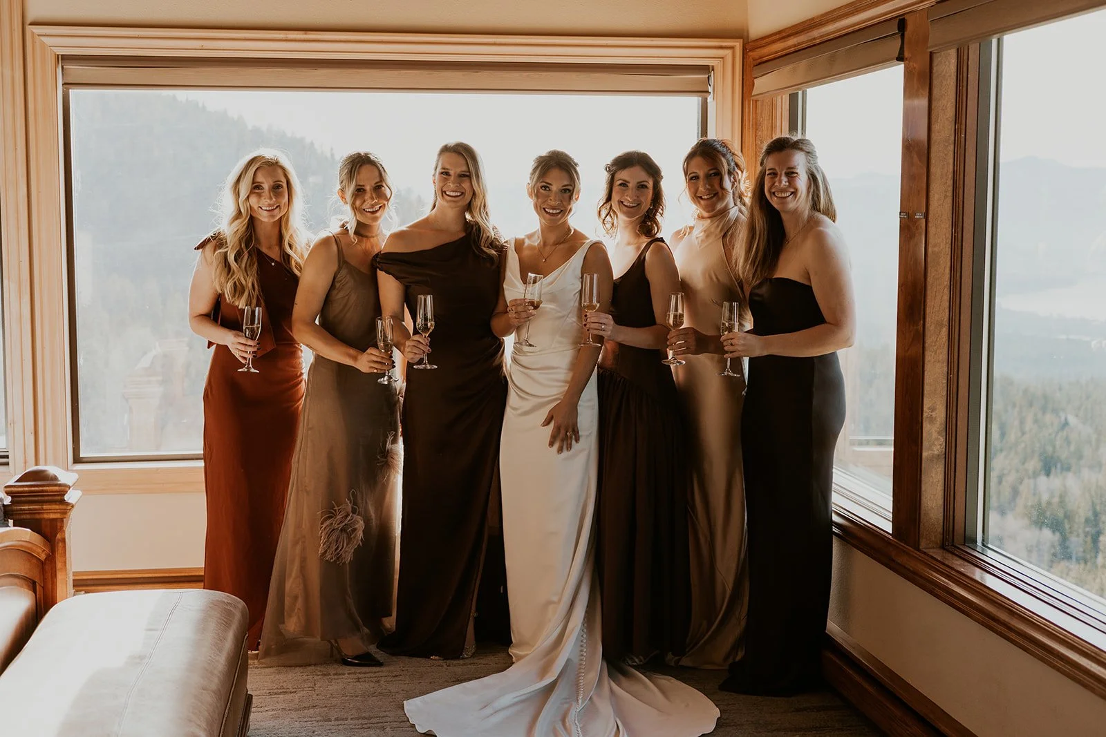 A bride in her wedding dress surrounded by her bridesmaids wearing brown dresses, champagne dresses, and rust dresses, holding champagne and smiling against the scenic windows of Tahoe Blue Estate.