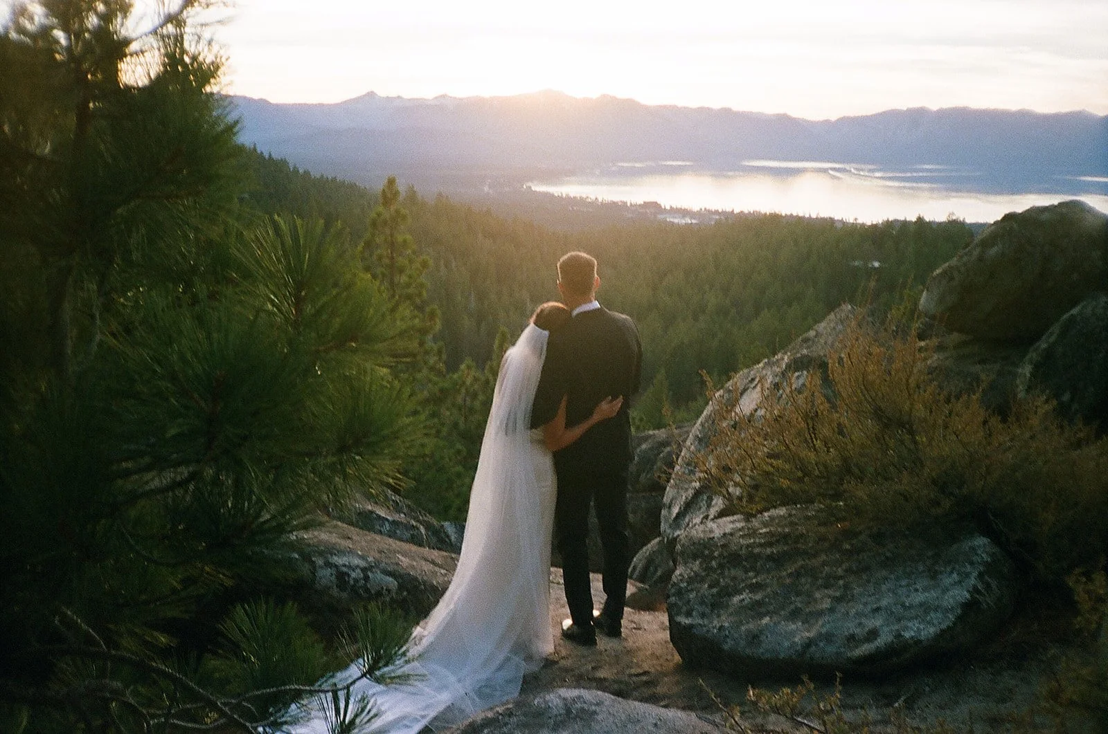 A film photo of the bride and groom embracing and standing on a rocky ledge looking at the sun setting over Lake Tahoe.