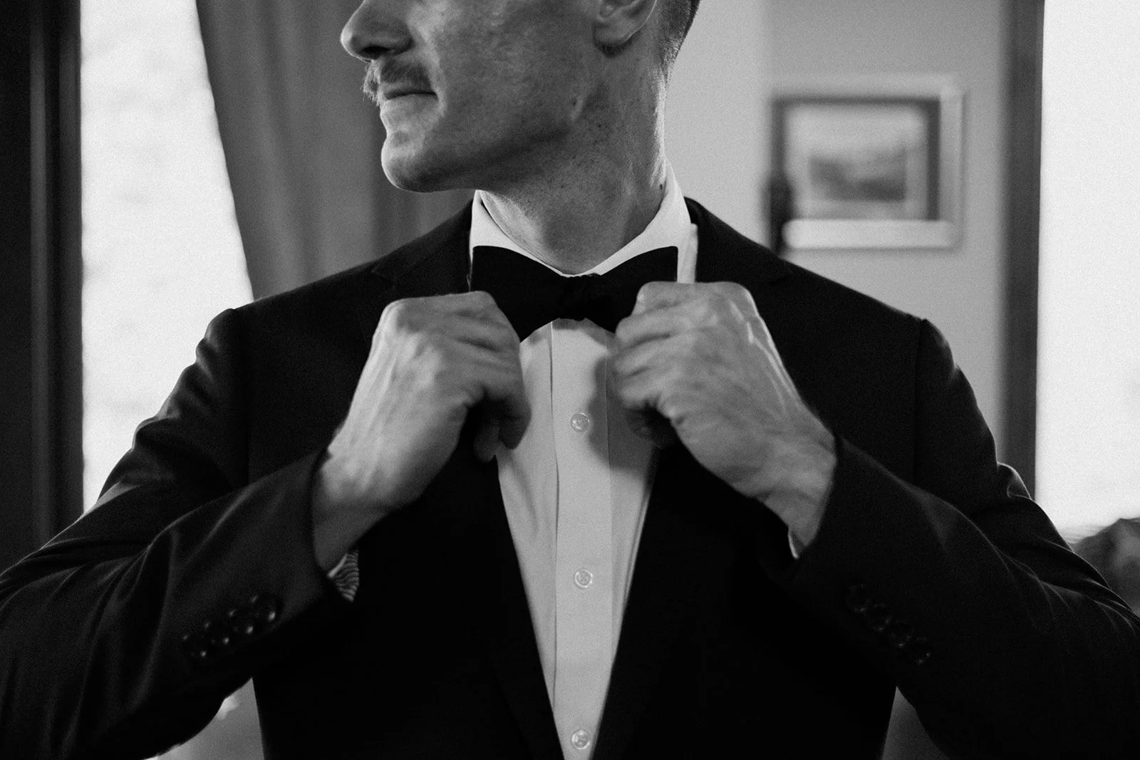 A black and white close up of the groom adjusting his tie and looking to the left.