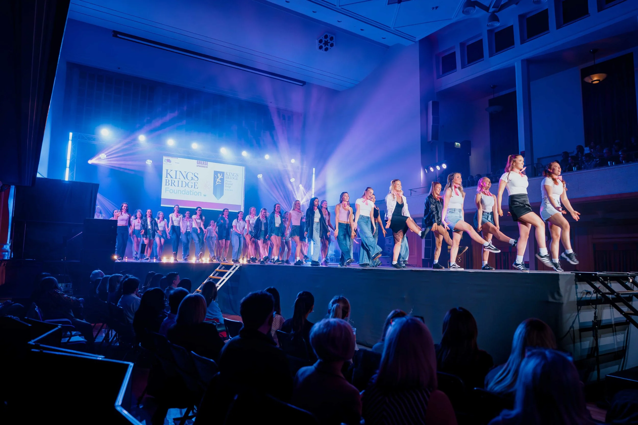 A group of young girls performing on stage at a school or community event, with an audience watching in front and a large screen displaying a logo for Kings Bridge Foundation in the background, with purple stage lighting.