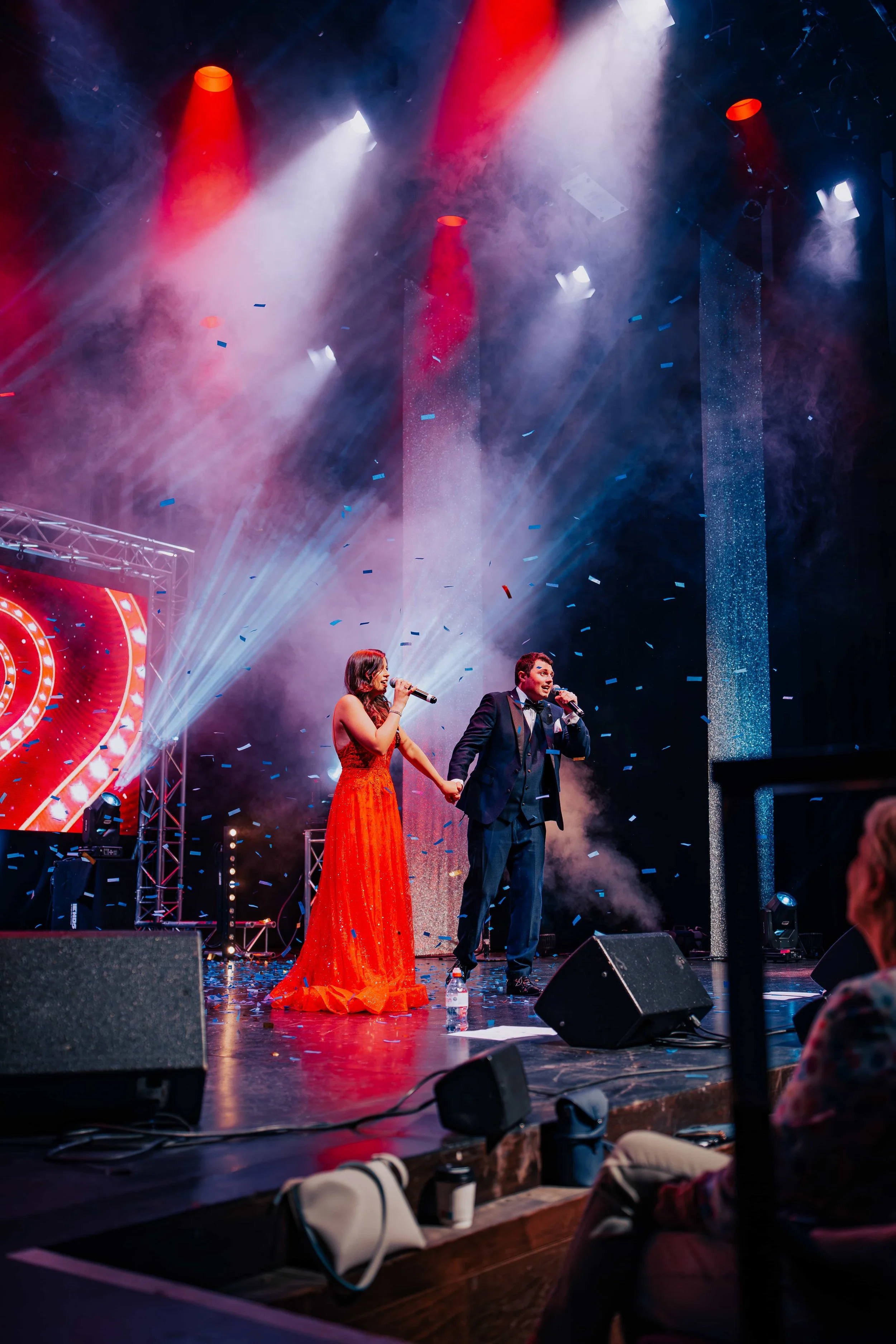 Male and female performers holding microphones on stage, with bright lights, confetti, and smoke effects during a concert or show.