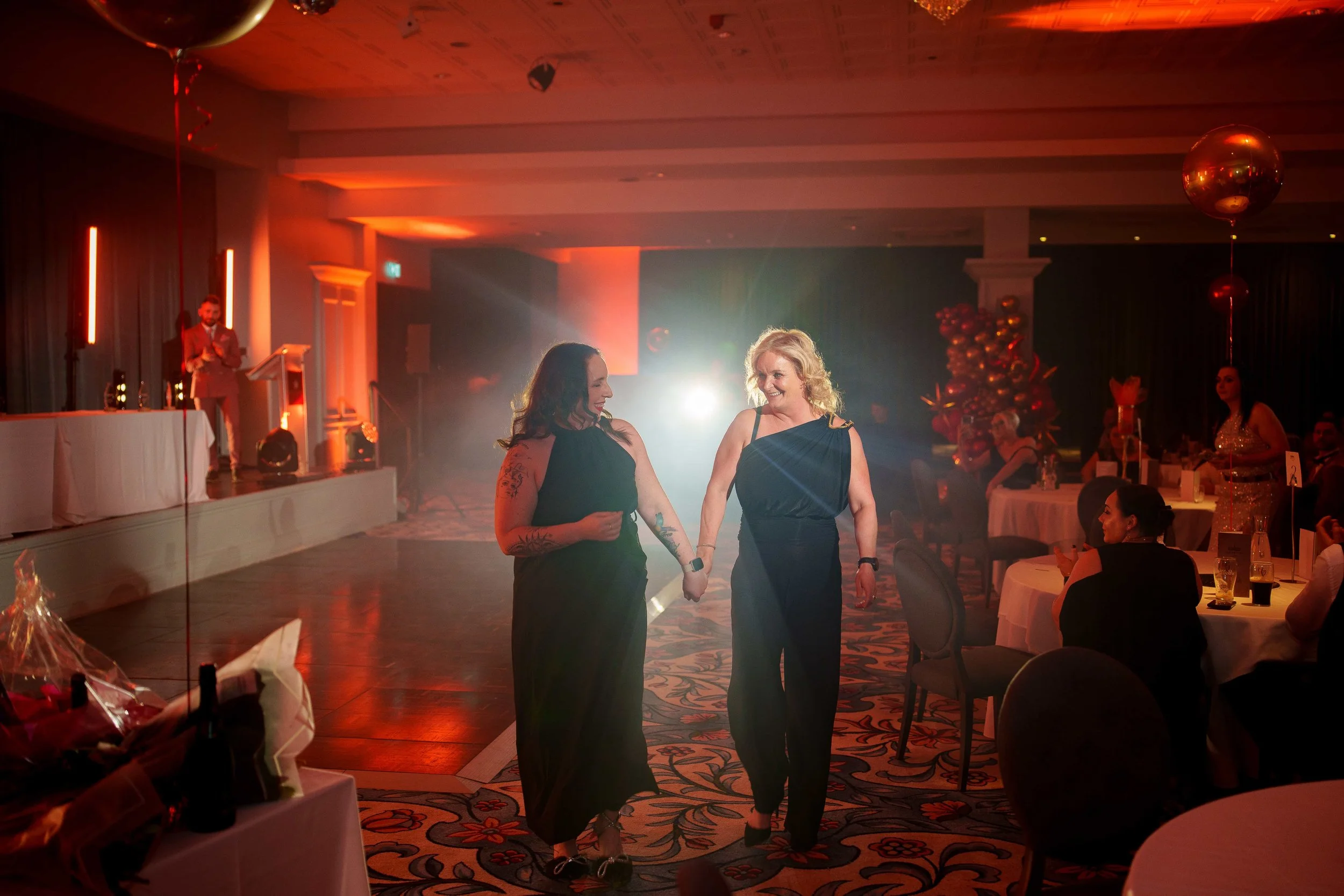 Two women dressed in formal attire dancing together at a banquet hall decorated with balloons and festive ornaments.