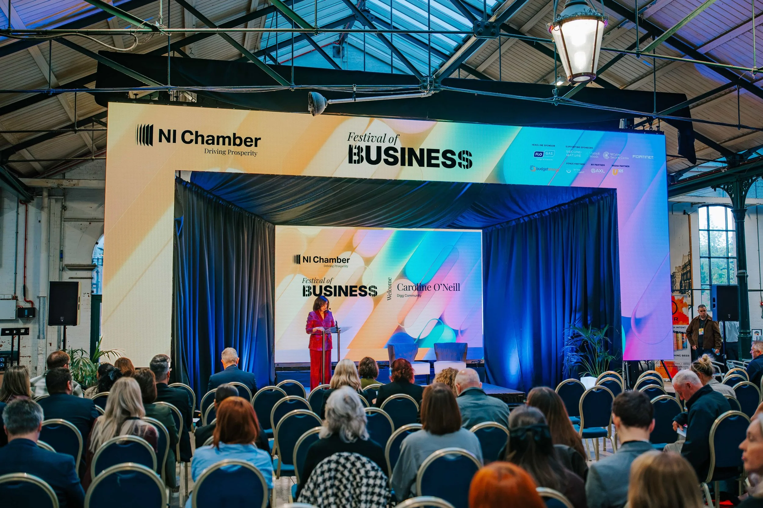 Host Caroline O'Neill at a glass lectern on the NI Chamber Festival of Business 2026 main stage at St George's Market, Belfast, framed by a 9.5m LED video wall arch with blue-lit drapes and a second LED wall behind the stage.