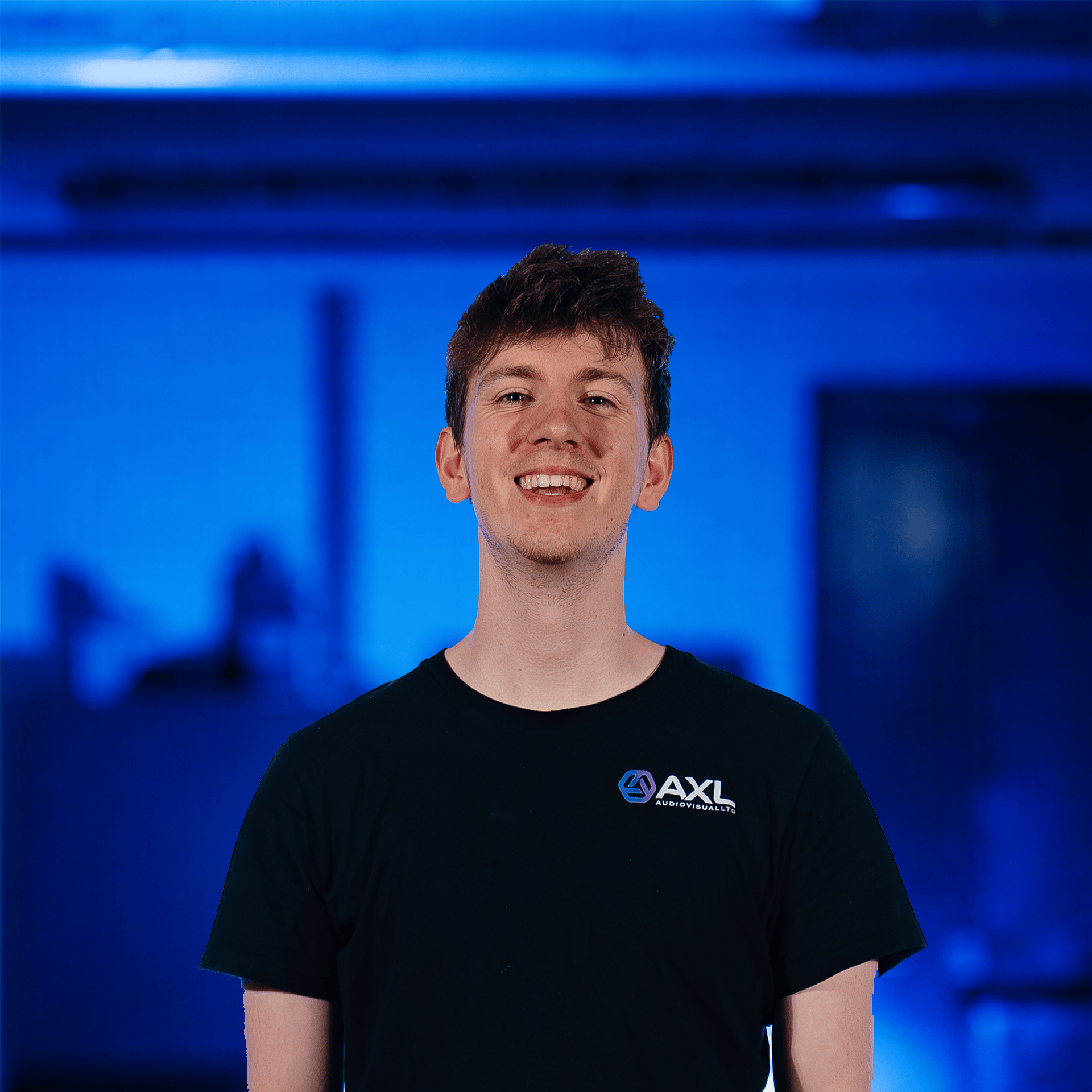 Young man with dark hair smiling, standing in front of a background with repeated blue logos, wearing a black T-shirt with a logo that reads 'AXL'.