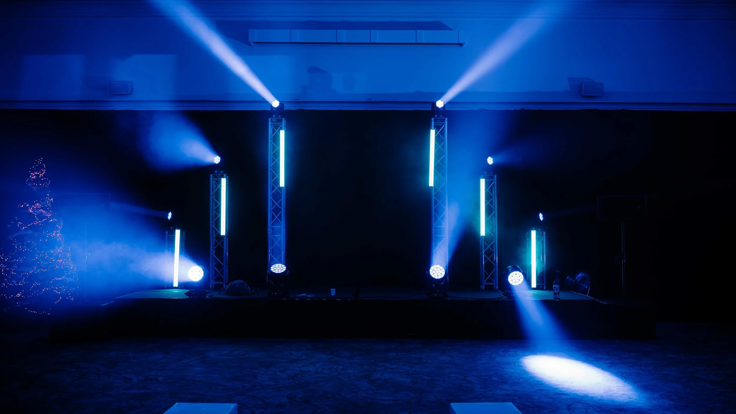 Stage with blue lighting, several vertical light fixtures, and a Christmas tree decorated with lights on the left side.