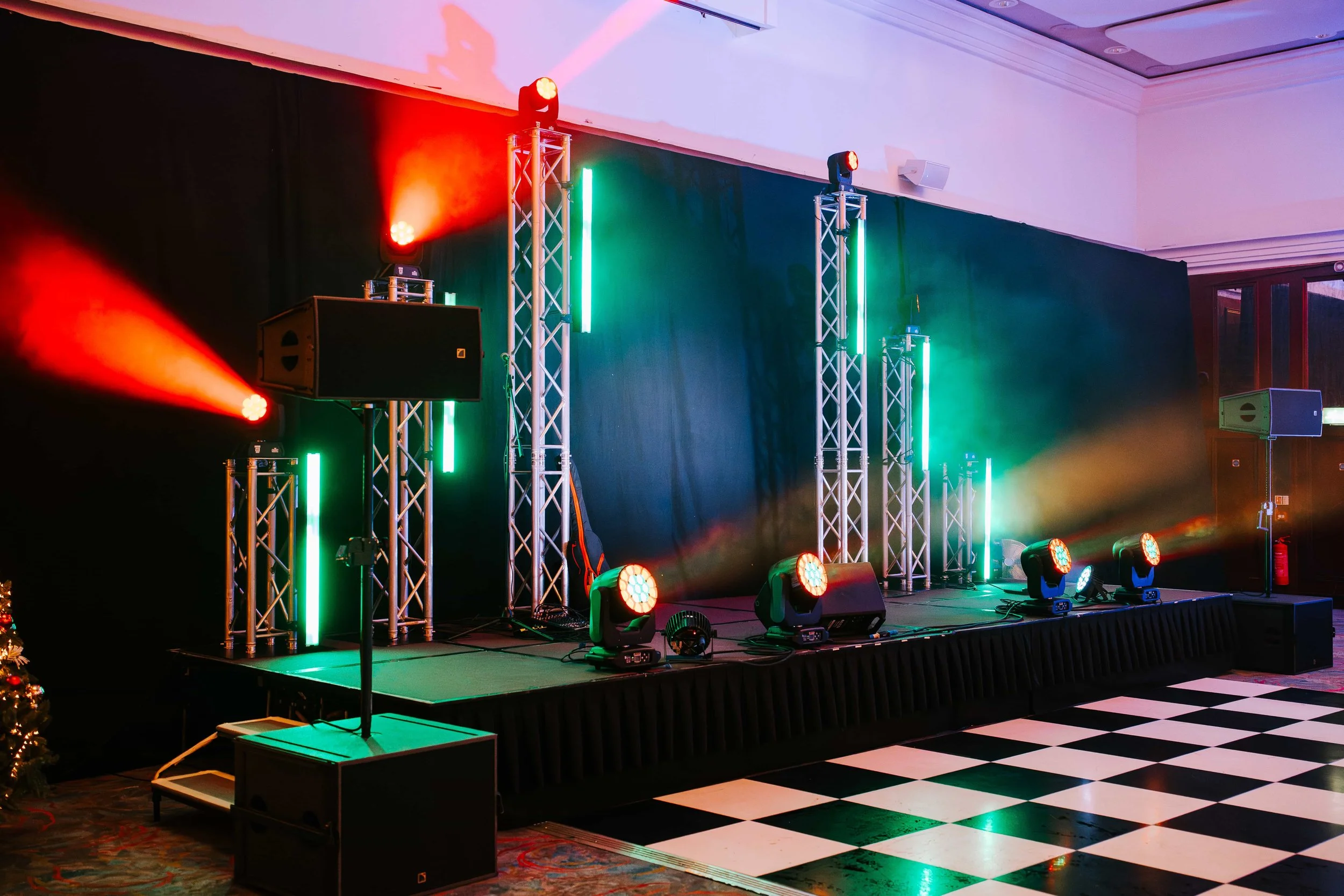 Event stage with colorful LED lights, speakers, and trusses set up for a performance, inside a venue with a black and white checkered floor.
