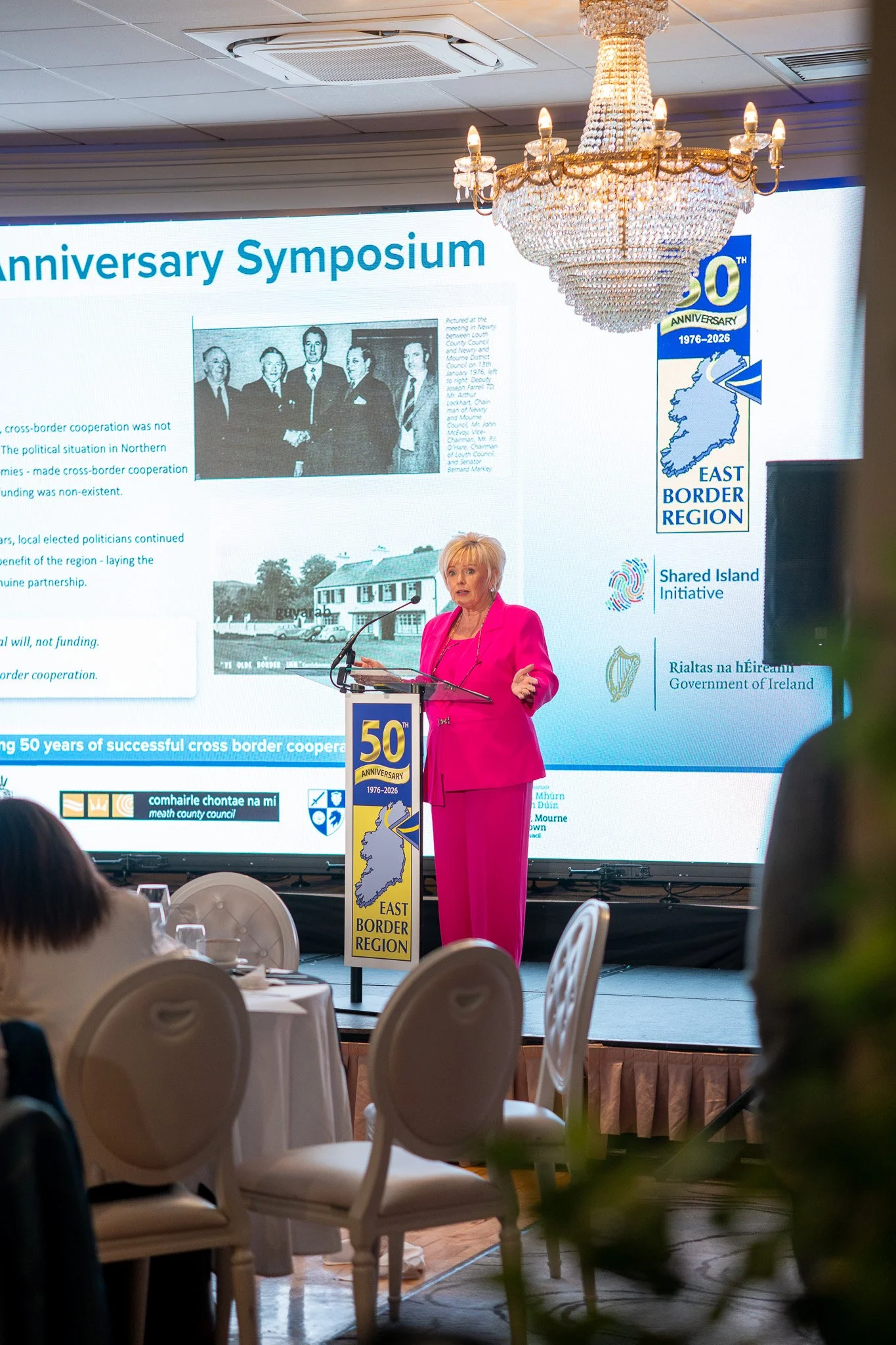 Speaker at a glass lectern with 50th anniversary branding, East Border Region history slide on the LED video wall behind.