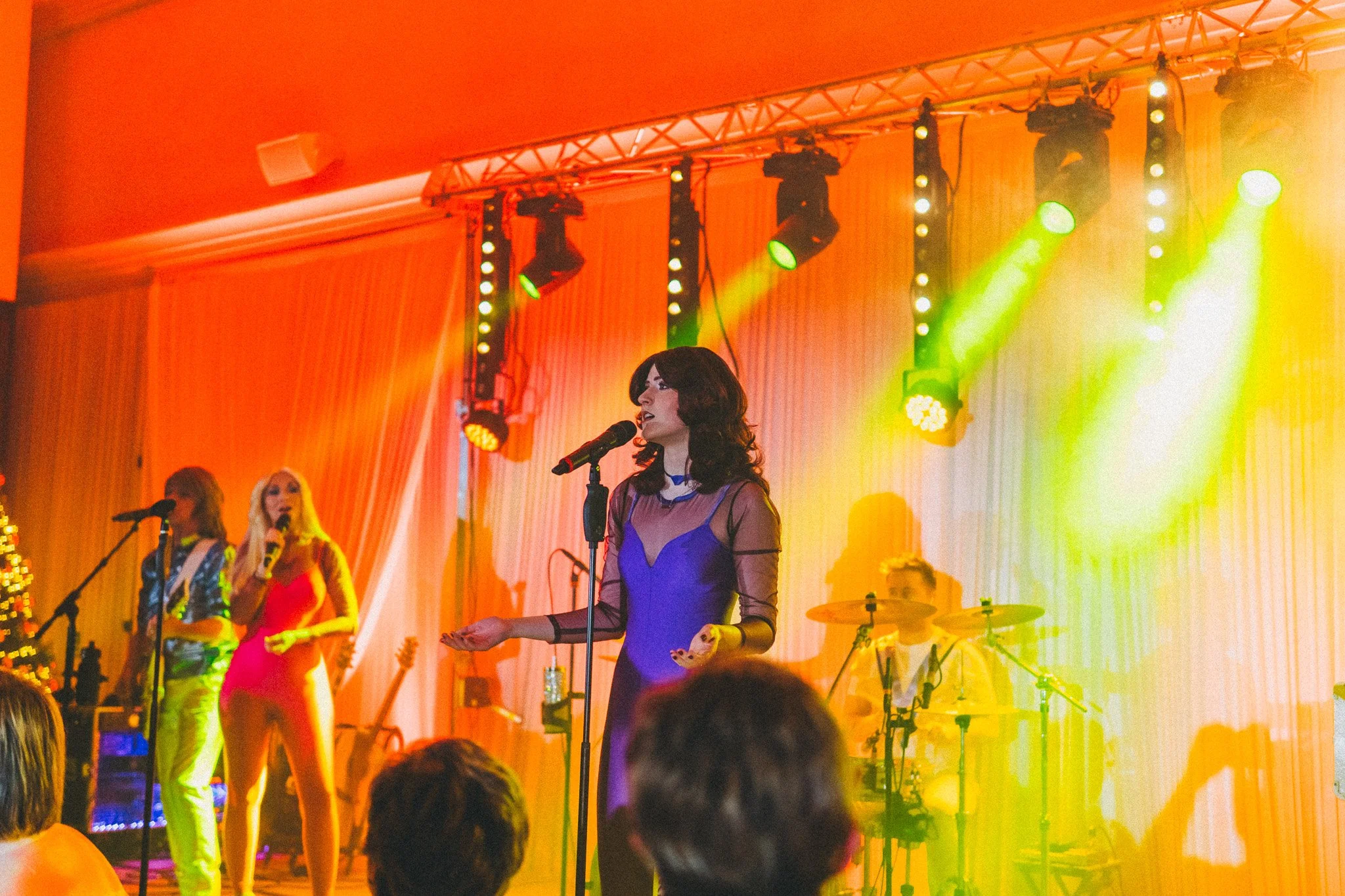 A woman in a blue dress singing into a microphone on stage with colorful lighting, backup singers, and musical instruments in the background.