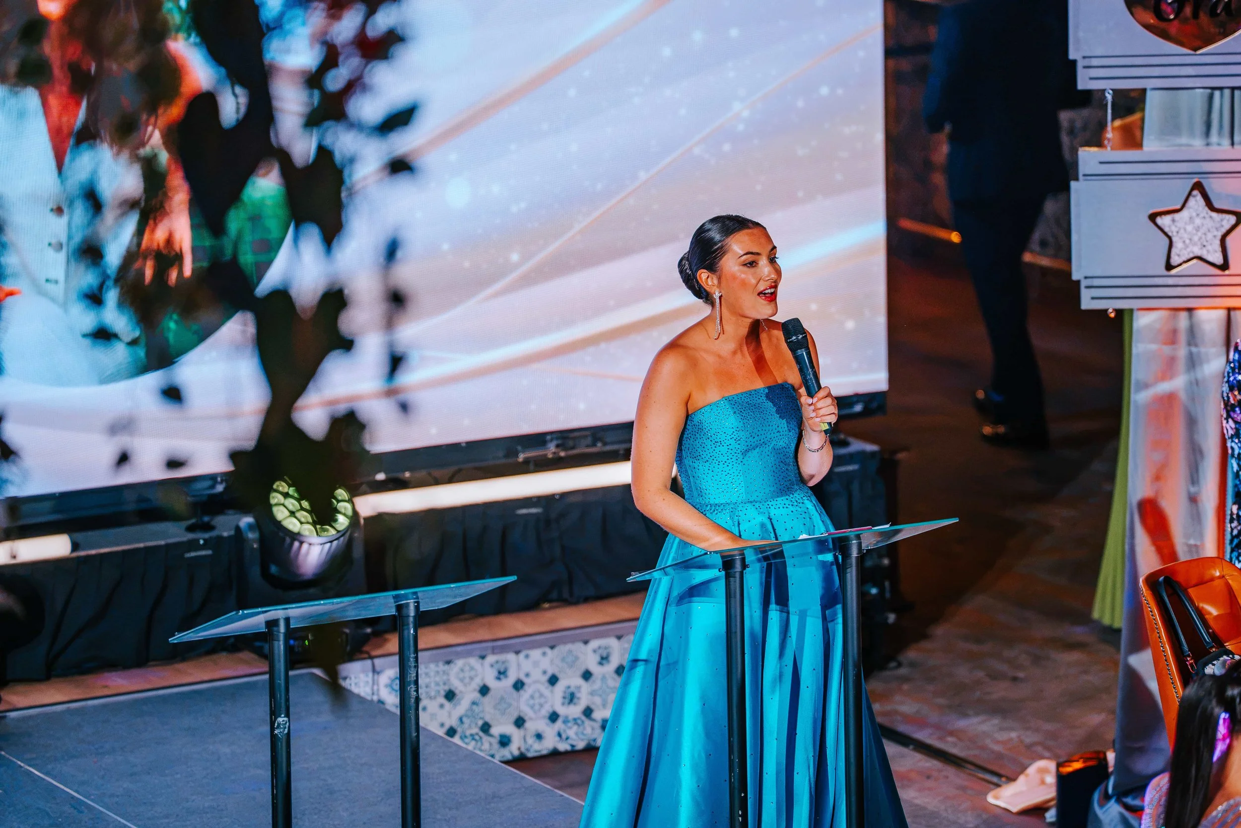 Awards host in a blue gown speaking at a glass lectern in front of the LED wall at the VAVA Influence Awards 2026