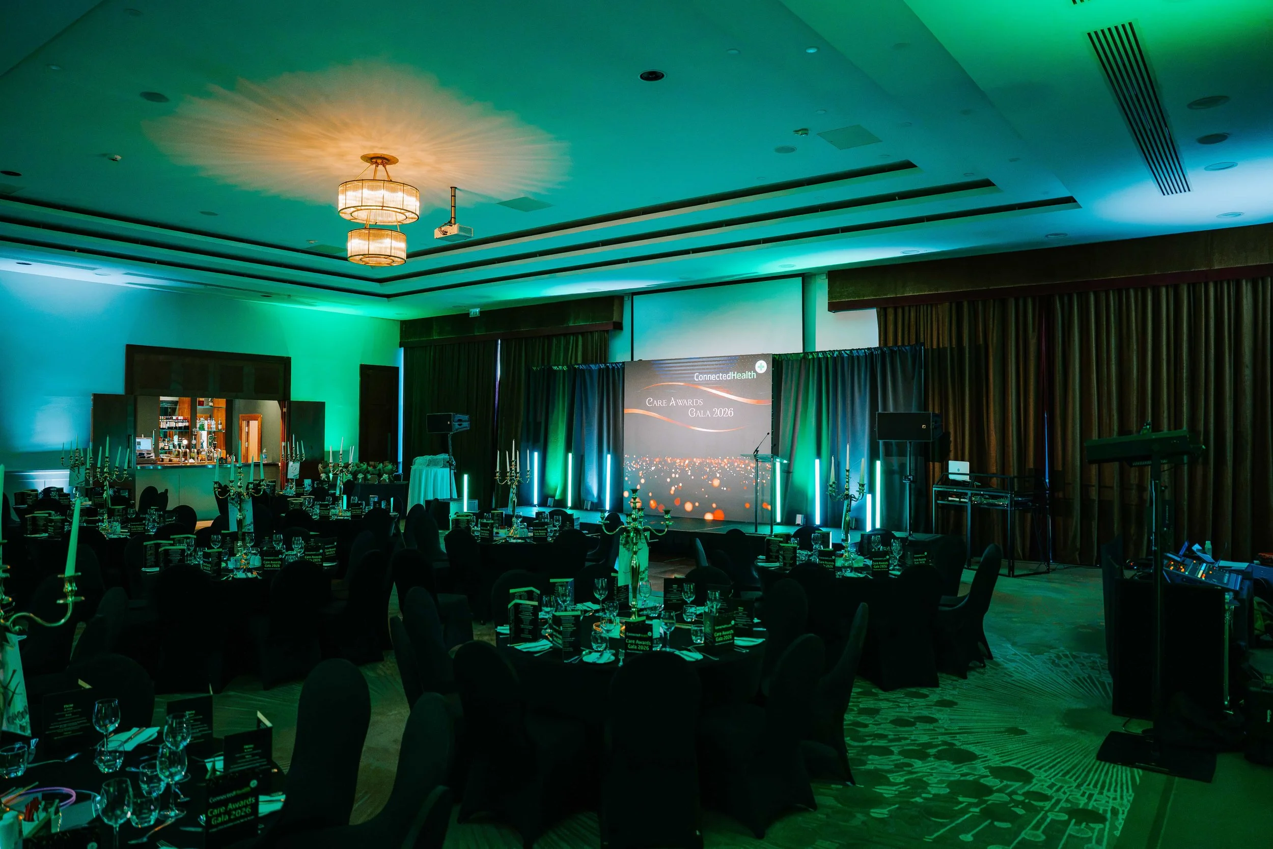 Awards dinner set with black-dressed round tables, gold candelabras and a central LED wall lit in green and teal