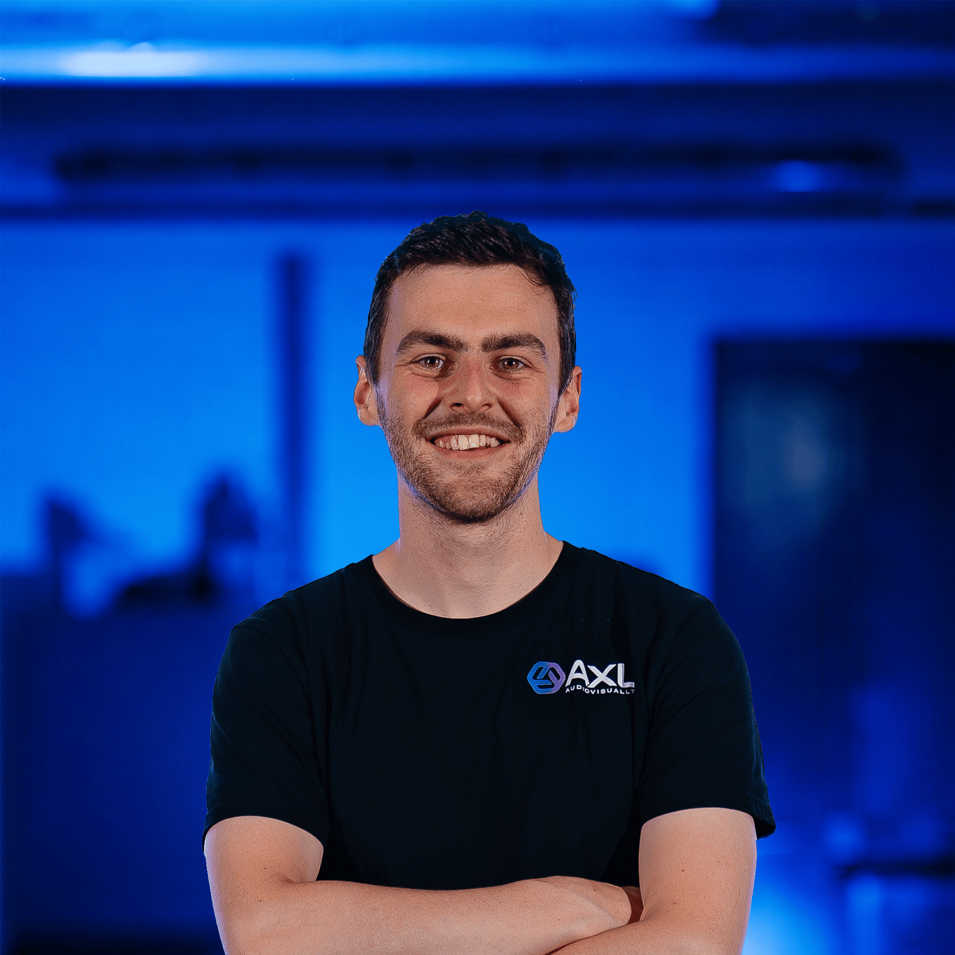 A young man with short dark hair and a beard, smiling and standing with his arms crossed, in front of a backdrop with repeating logo patterns. He is wearing a black shirt with a logo that says 'AXL' on it.