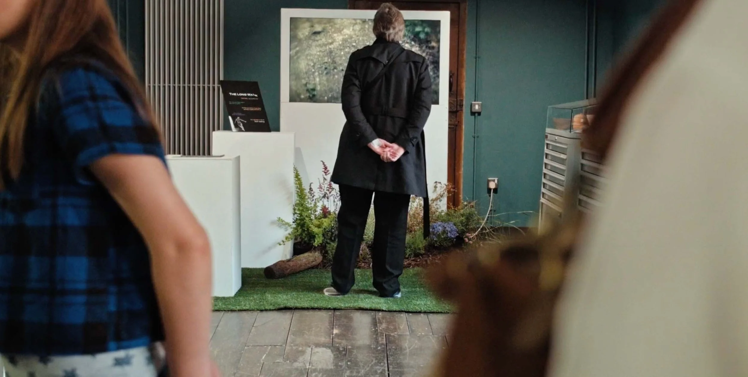 A person walking on green grass in front of a screen with plants around them, all set in a museum.