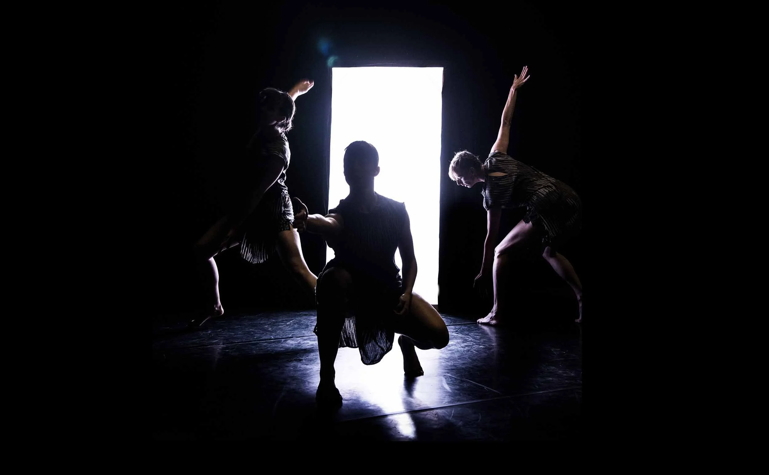Silhouette of three female dancers in different dramatic poses, backlit by a white box of light.