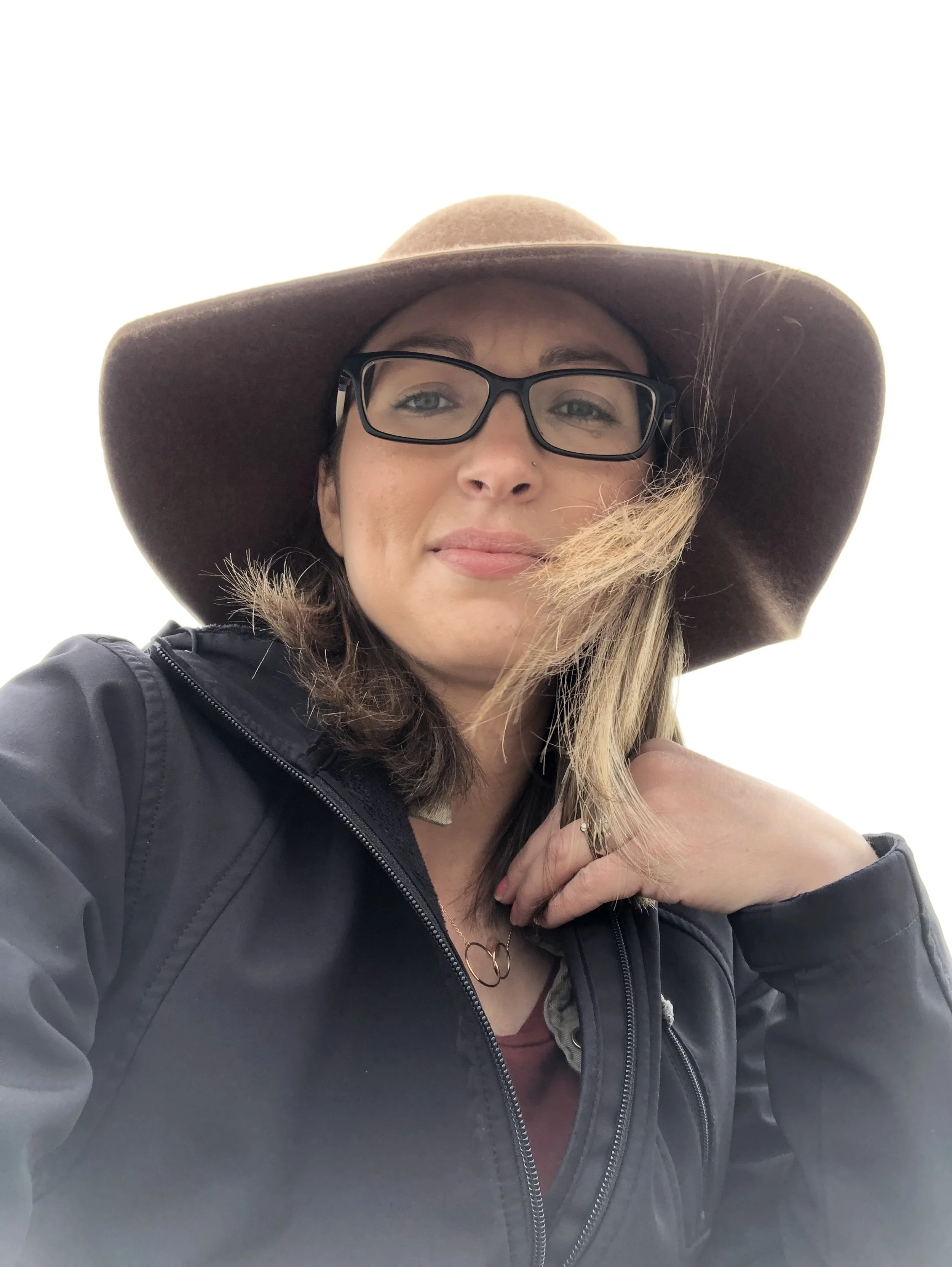 A woman wearing glasses, a large floppy hat, and a black jacket, with light brown hair and a subtle smile.