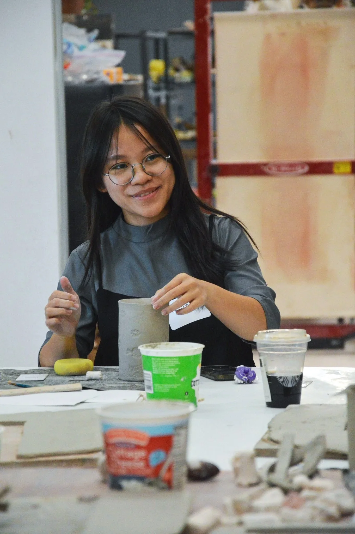 A young woman wearing a name tag looks over with a smile as she works on her handbuilt clay mug