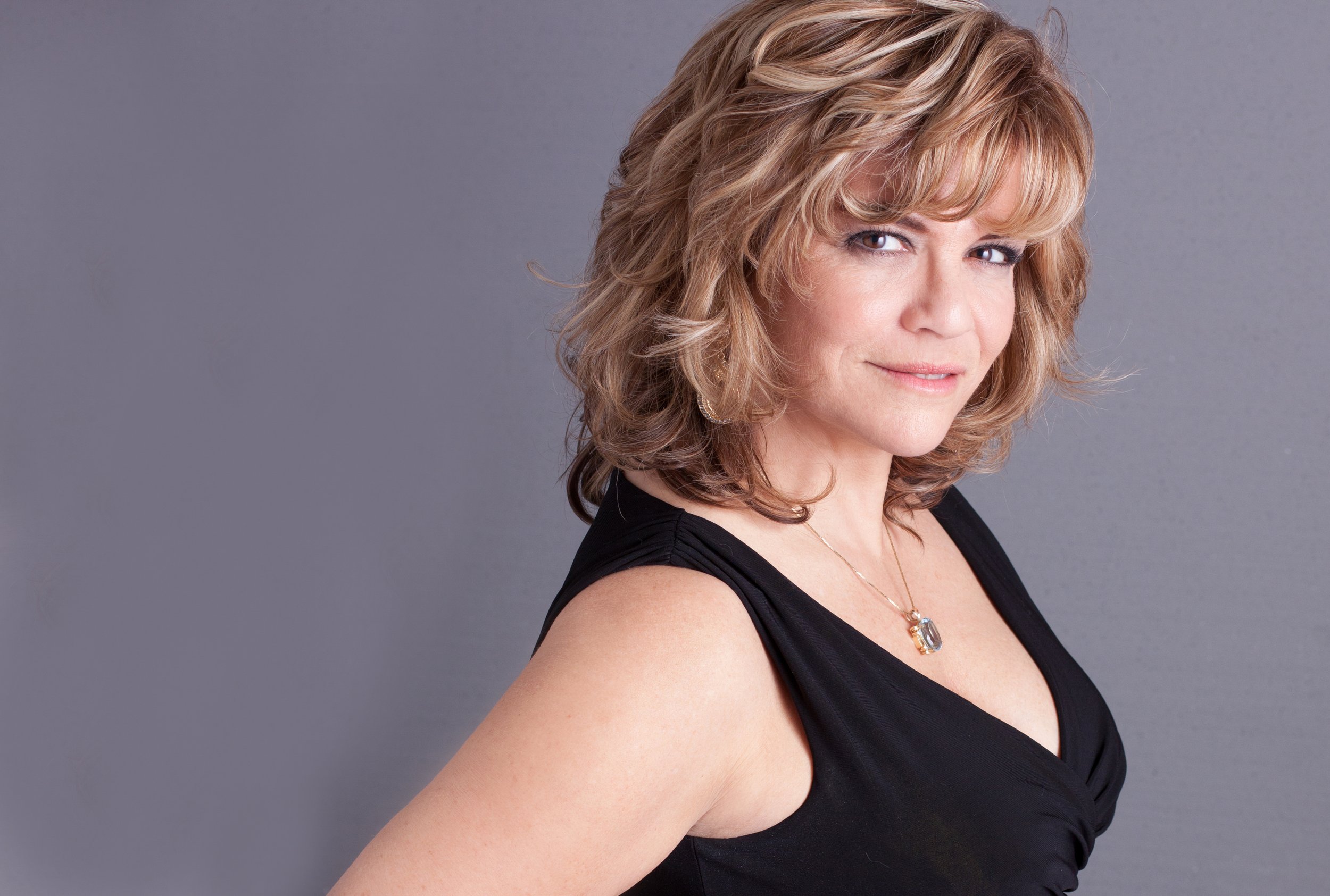 Woman with wavy blonde hair in a black sleeveless top, wearing a necklace, posing against a gray background.