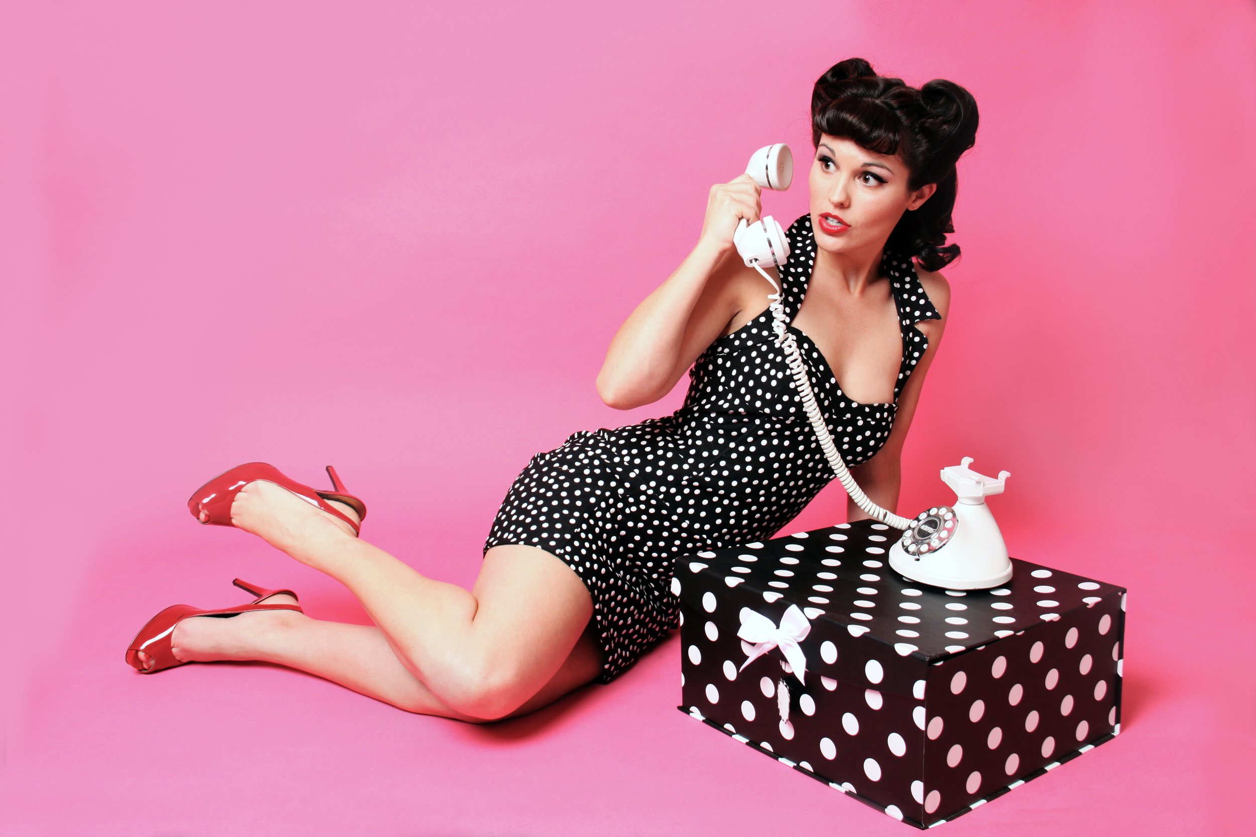 A woman in a polka dot dress posing with a vintage phone against a pink background.