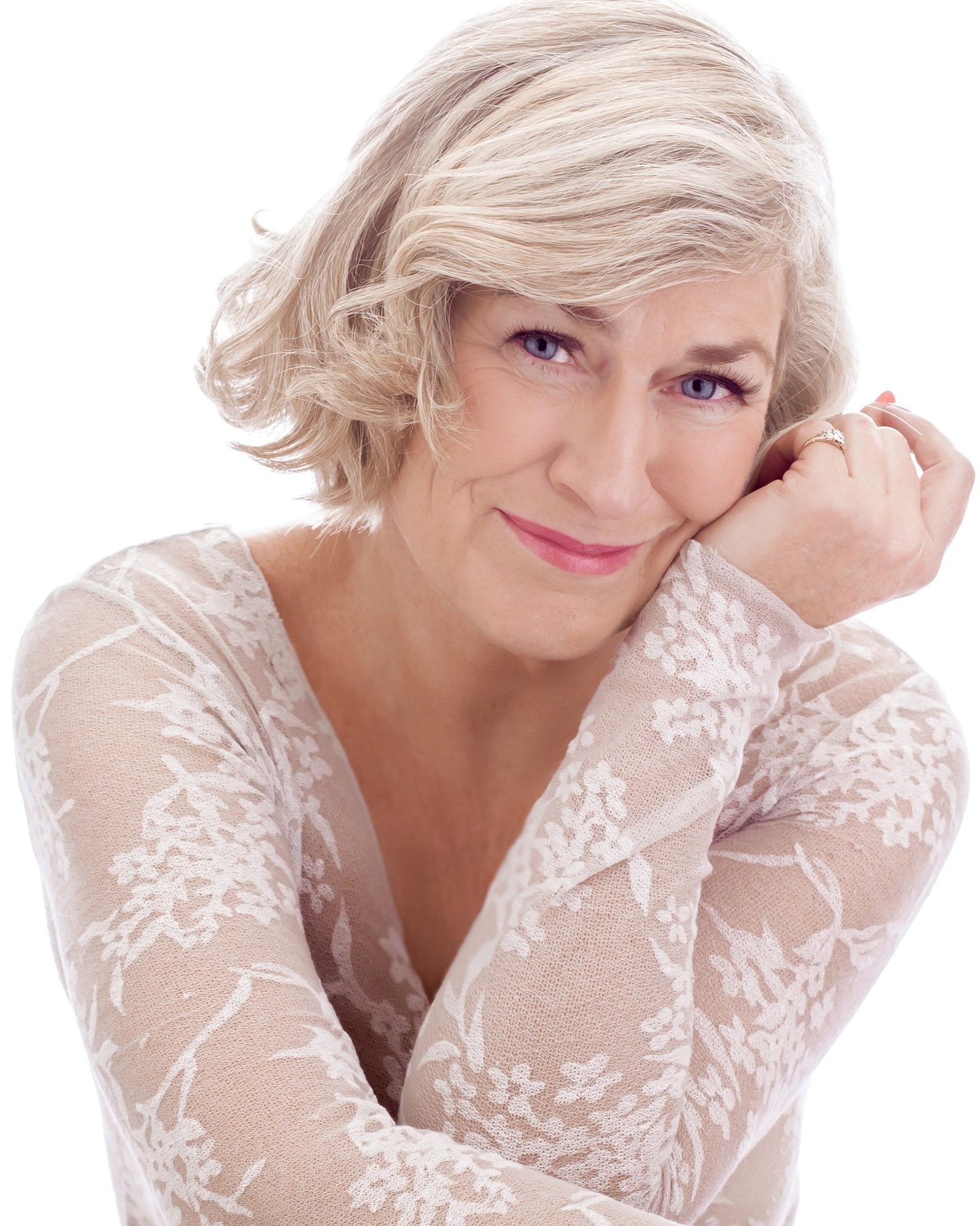 Senior woman with light hair wearing a floral lace top, smiling and resting her head on her hand.