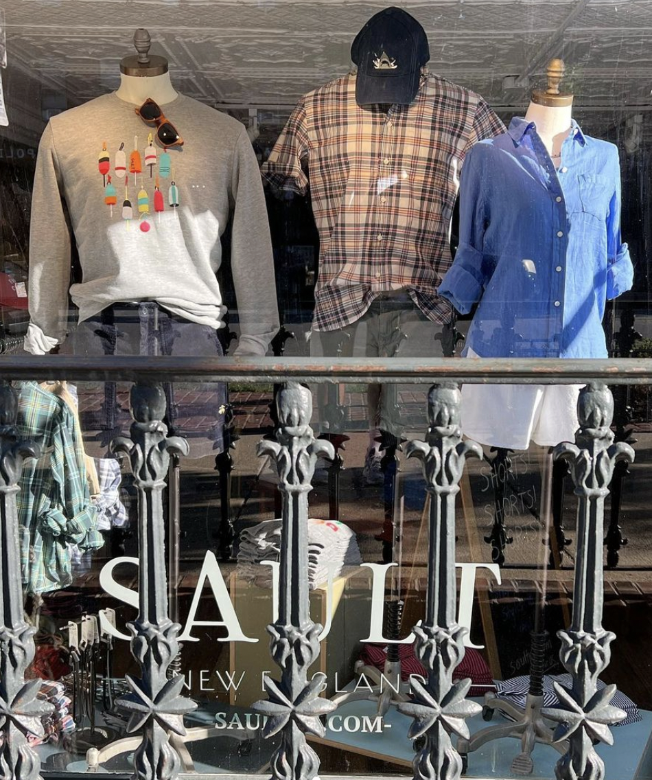 Storefront display with three mannequins dressed in casual clothing: a gray sweatshirt with prints and sunglasses, a plaid shirt with a black cap, and a blue button-up shirt with rolled sleeves. Text on the window reads 'SAULT NEW ENGLAND' with websi