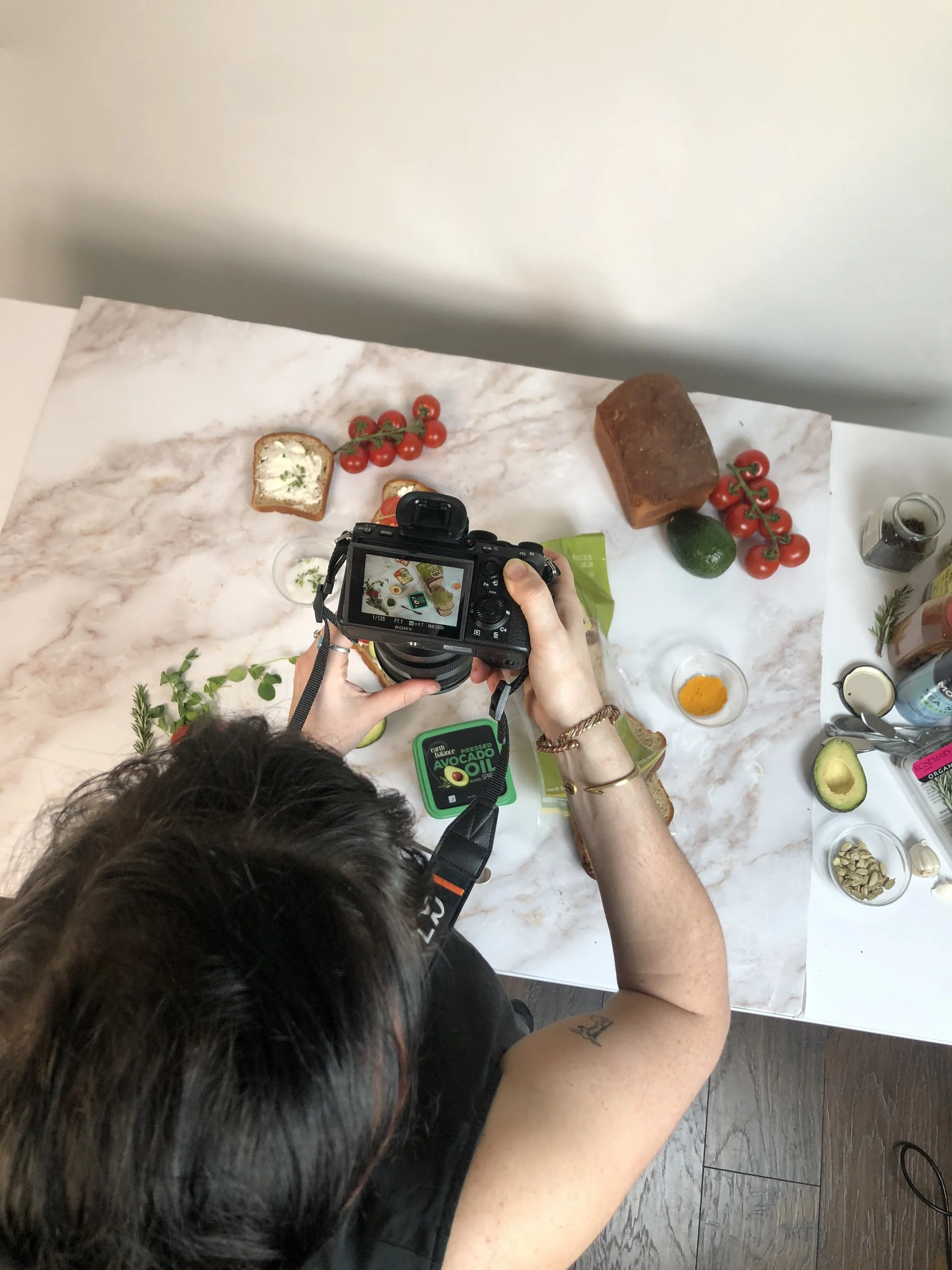 Overhead view of a person photographing food items on a marble surface. Included are a loaf of bread, slices of bread with spread, cherry tomatoes, an avocado, herbs, a small bowl of turmeric, seeds, and a container of avocado oil.