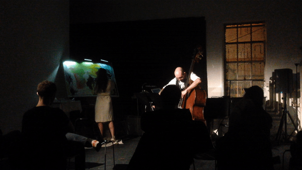 A dimly lit stage with a woman painting on a canvas under a bright light and a man playing a double bass. Several audience members are seated, observing the performance.