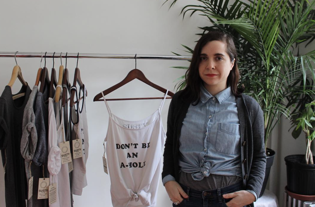 Woman standing next to a clothing rack with assorted tops on hangers. A white tank top with a slogan is prominently displayed. There are potted plants in the background.