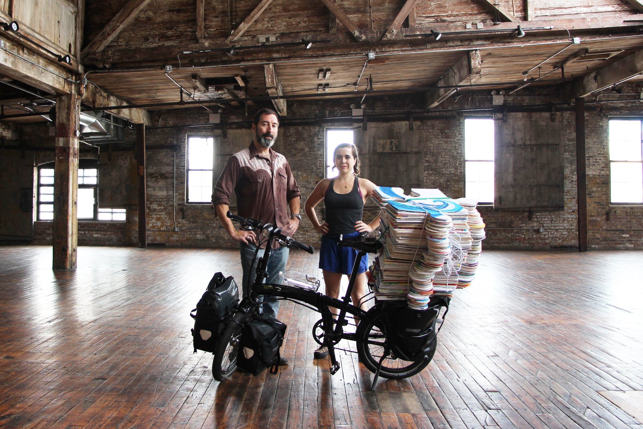 Two people standing next to a bicycle loaded with stacks of books in a spacious, rustic warehouse.