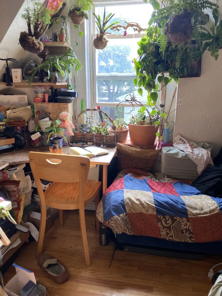 View of a bedroom with a desk, window, and bed. There are many plants, art materials, other other ephemera on the desk next to the bed. The bed has a white, red, and blue patchwork quilt.