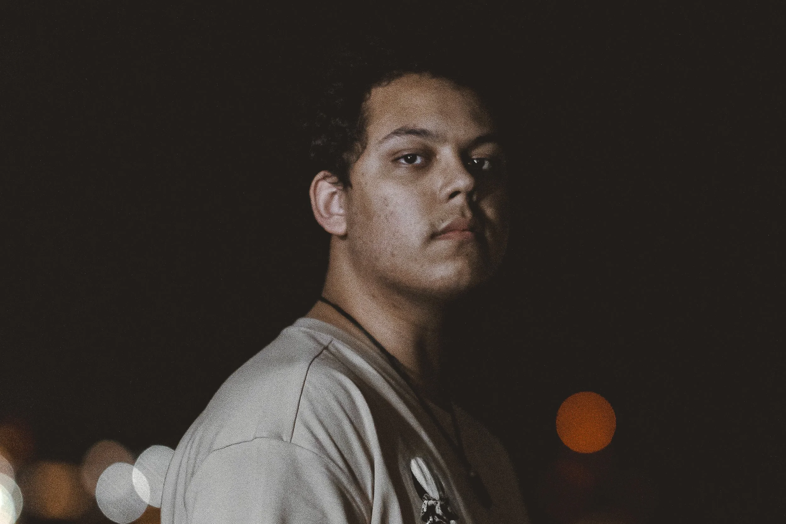 A photograph of a young dark skinned person looking directly at the camera with a lens flare on their right shoulder