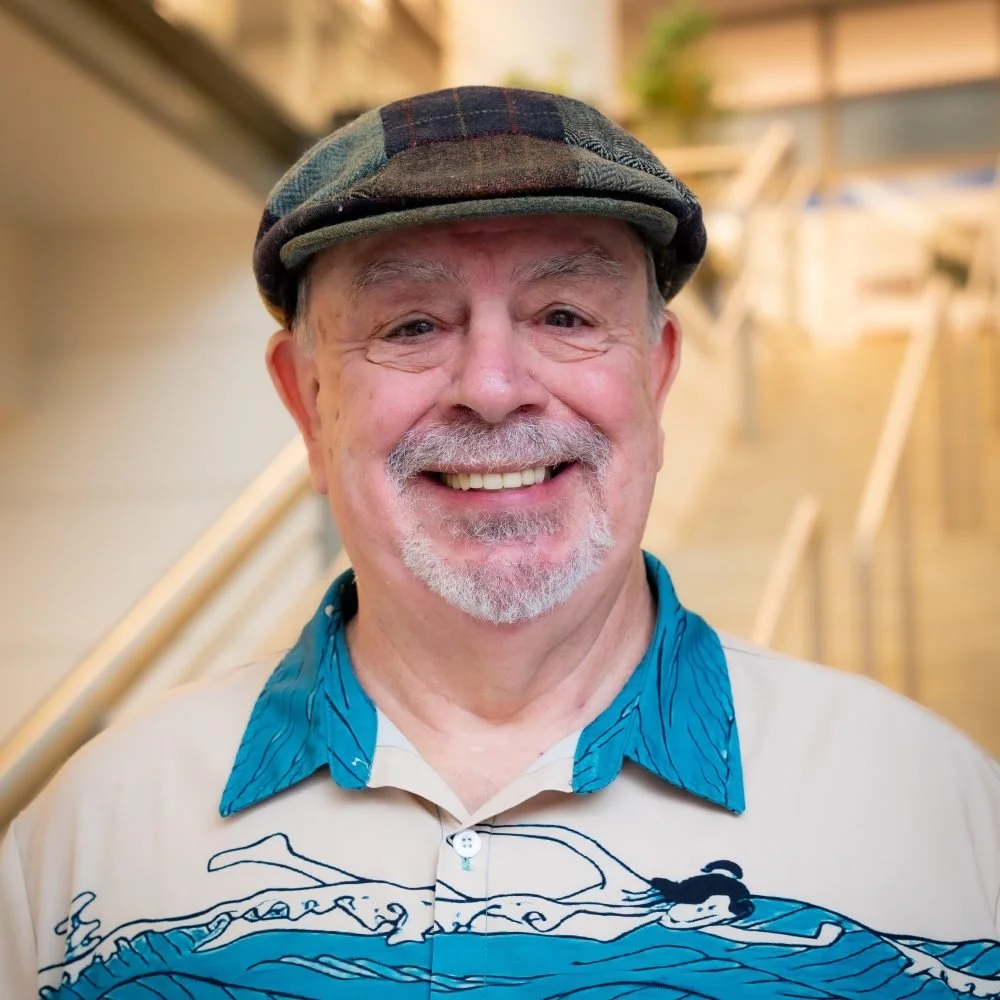 Smiling elderly man wearing a plaid cap, beige shirt with blue ocean wave design, and a blue collar, standing in an indoor space with wooden stairs in the background.