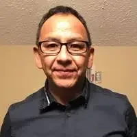 A man with glasses and short dark hair smiling, wearing a dark button-up shirt, standing indoors against a light-colored wall.