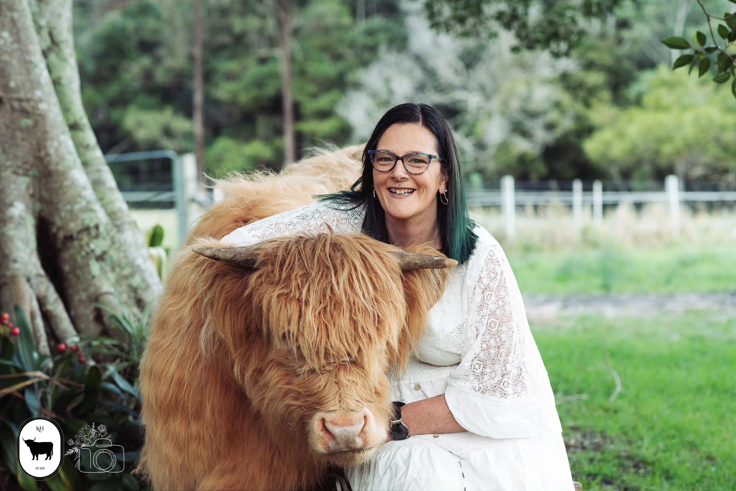 A woman with black hair and glasses smiling while cuddling a Highland cow outdoors in a green, wooded area.