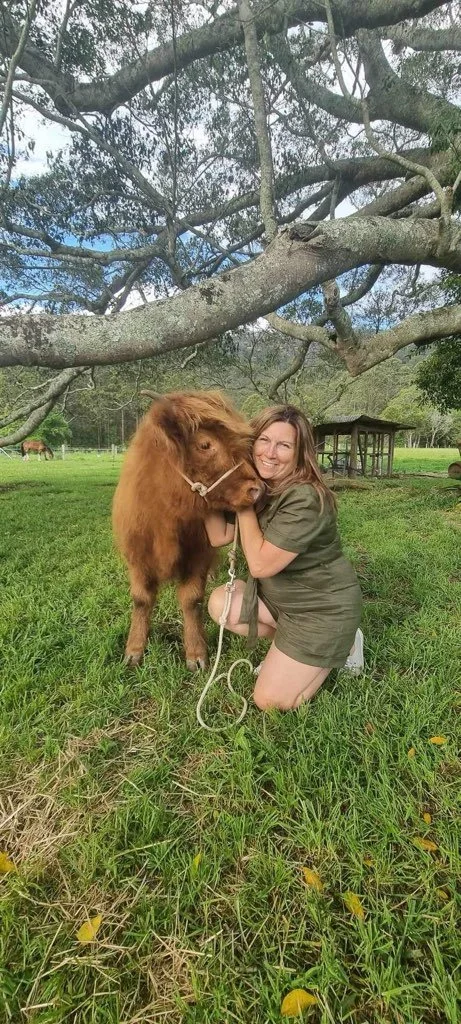A woman kneeling on the grass, hugging a brown cow, under a large tree with sprawling branches, in a rural setting with a small shelter and other cows in the background.