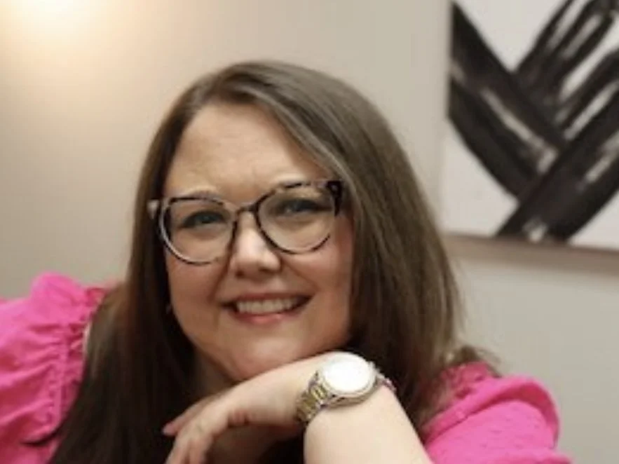 Smiling woman with glasses wearing a pink top, resting her chin on her hand.