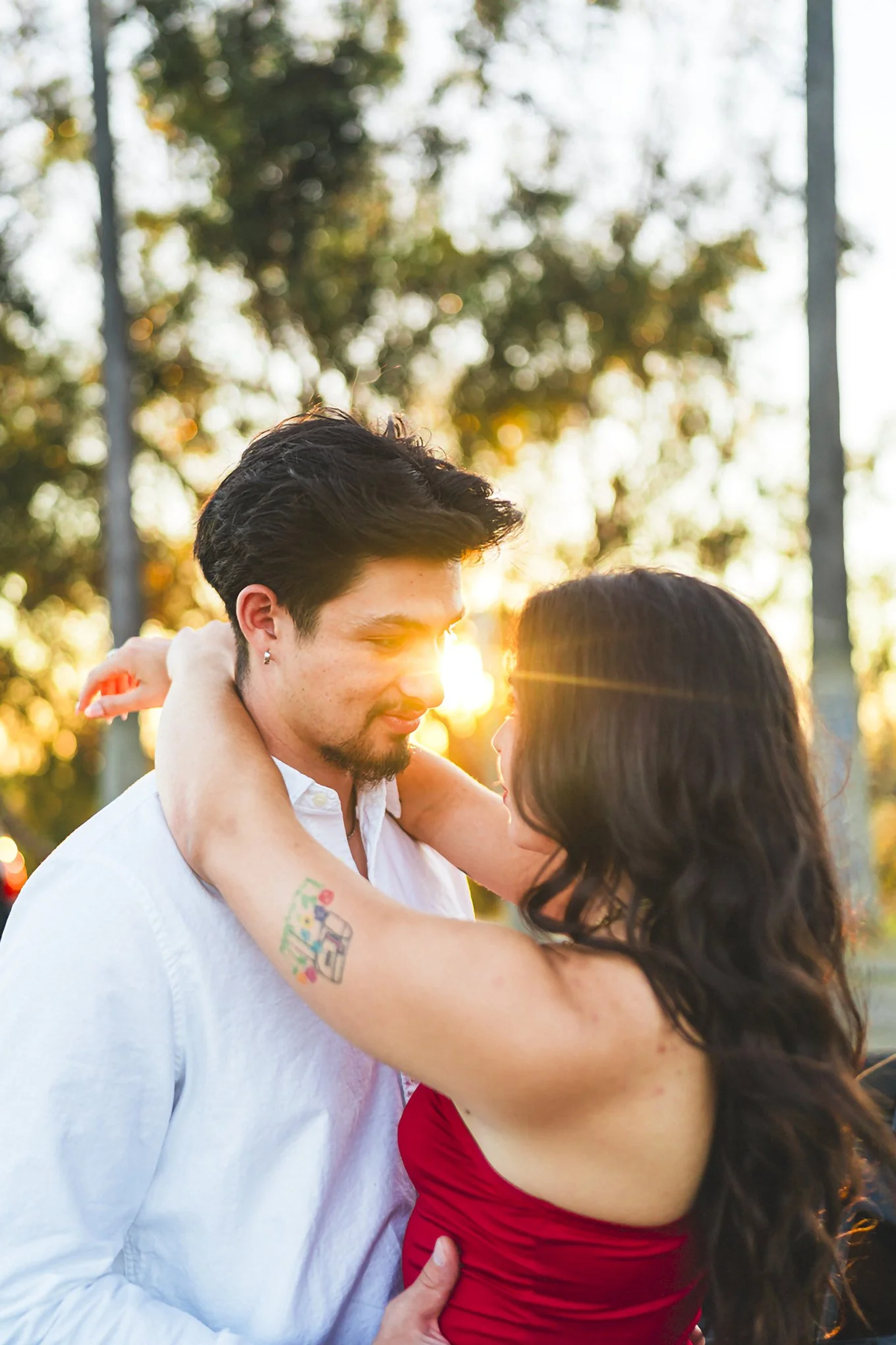 A romantic couple embraces during sunset, with warm sunlight illuminating their faces and a background of trees.