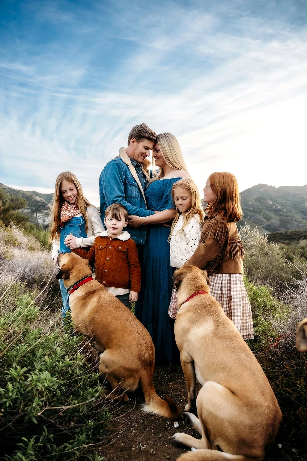 Family of five, including two dogs, standing outdoors in a natural landscape with green hills and blue sky.