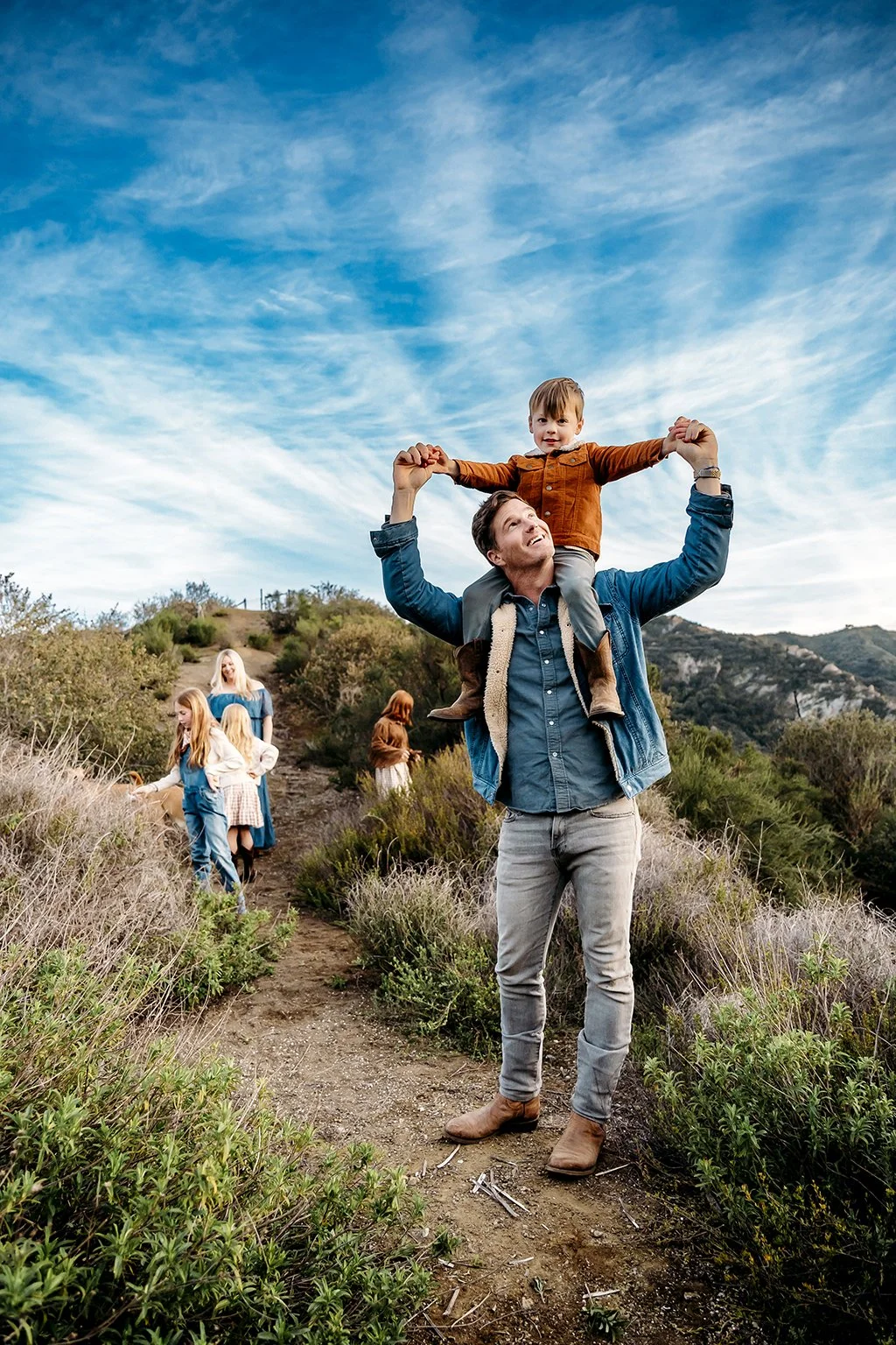 A man carrying a young child on his shoulders while walking on a trail in a hilly, green landscape, with a group of women and children in the background under a blue sky with wispy clouds.