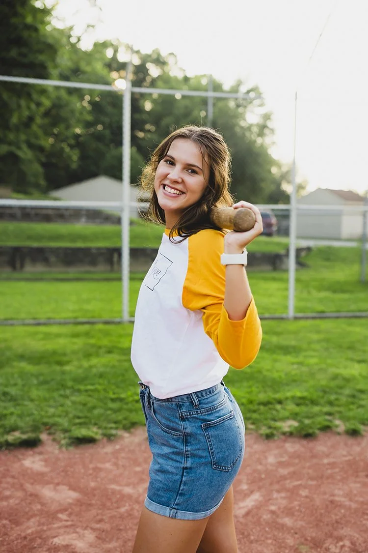 High school senior standing on a baseball field holding a bat, wearing a white and yellow jersey and denim shorts during a hobby senior portrait session in Hagerstown, Indiana.