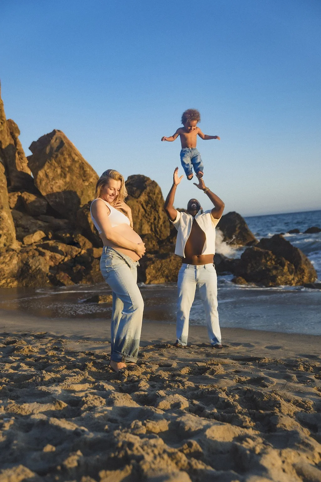 Family enjoying a beach maternity session with an expectant mother, father playing with their child, and joyful sunset moments during a destination lifestyle photography session by an Indiana family photographer.