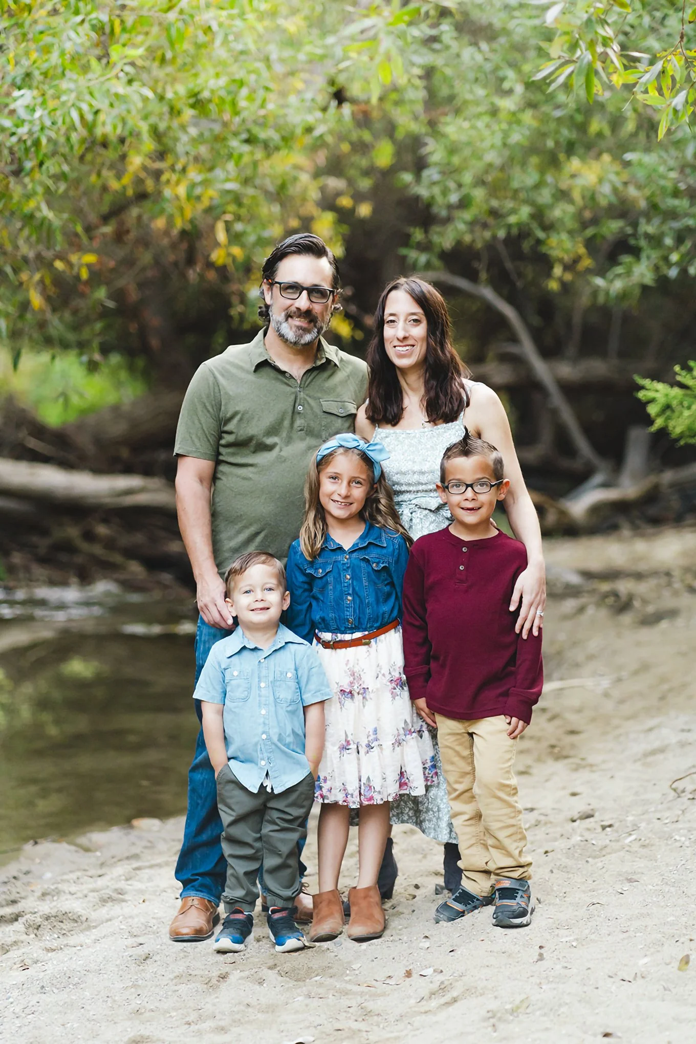 A Midwest family of five standing together outdoors near a creek, with trees and greenery in the background, smiling at the camera.