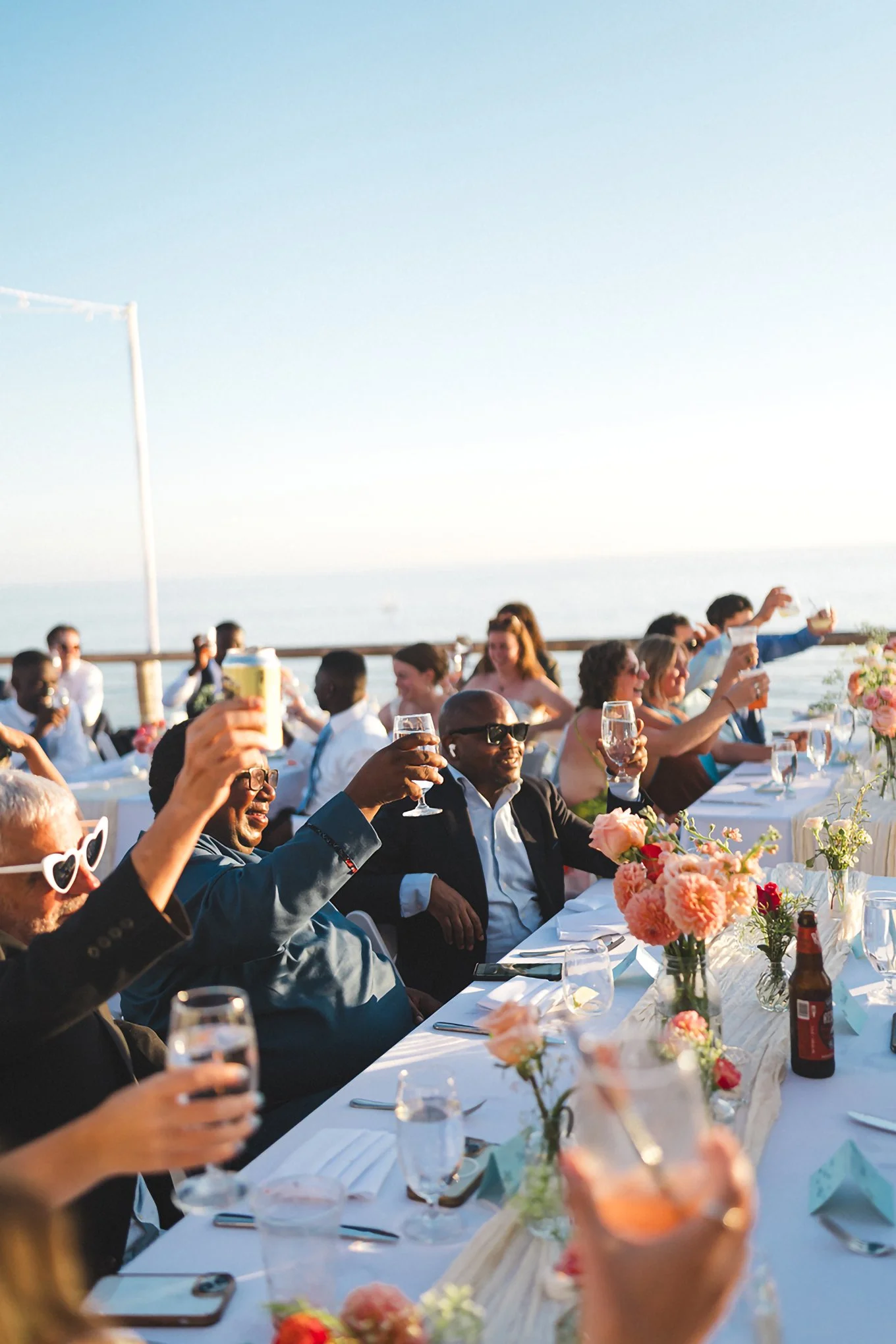 Intimate destination wedding by the water, guests raising glasses in a toast, outdoor wedding photography, story-driven celebration.