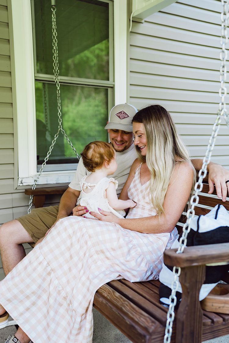 Parents holding their baby girl close while relaxing together on their front porch swing at home.