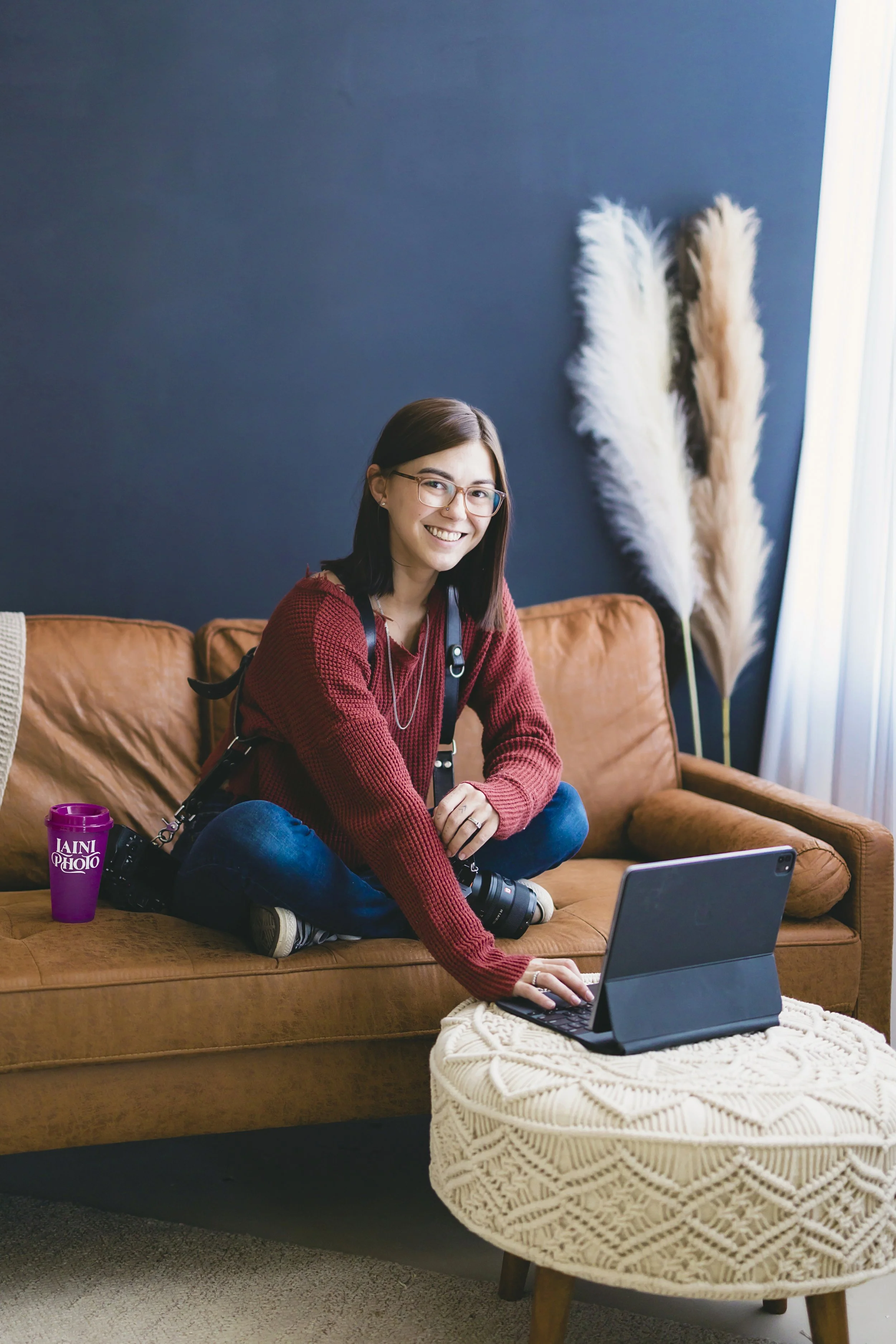 Richmond lifestyle photographer headshot: woman with camera on neck, using tablet, smiling on brown sofa with pampas grass.