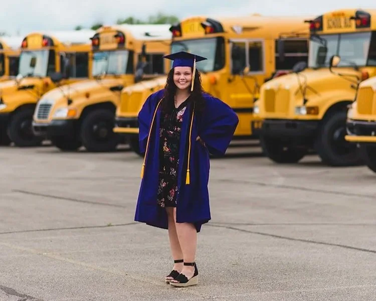 Indiana graduating Senior in a graduation gown and cap standing on pavement in front of yellow school buses, smiling.