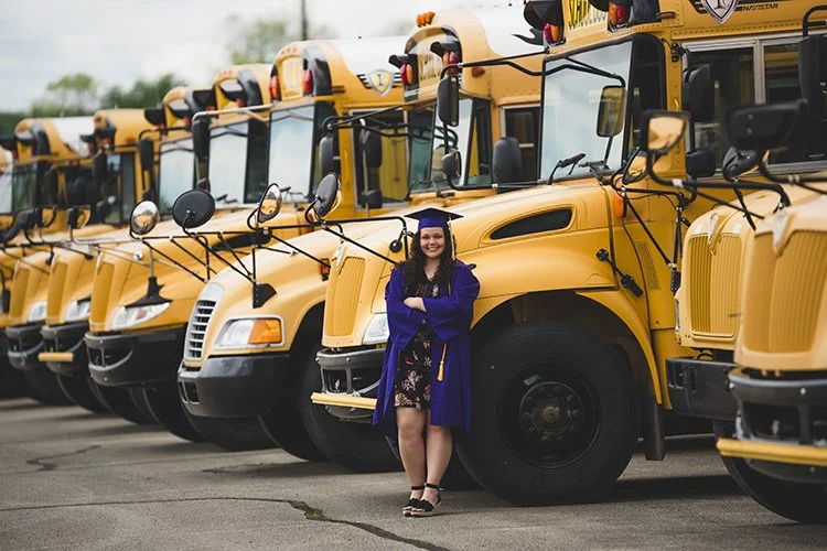 A graduating senior from Central Indiana in a graduation gown and cap standing in front of a row of yellow school buses.
