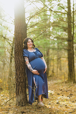 Pregnant woman with tattooed arms standing in a forest in Indiana, leaning against a tree in a blue dress and smiling.