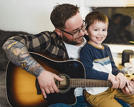 Indiana family photographer capturing father with tattoos playing guitar while holding smiling son in relaxed in-home session