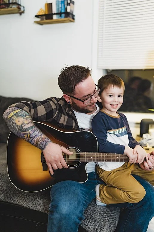 Indiana family photographer capturing father with tattoos playing guitar while holding smiling son in relaxed in-home session