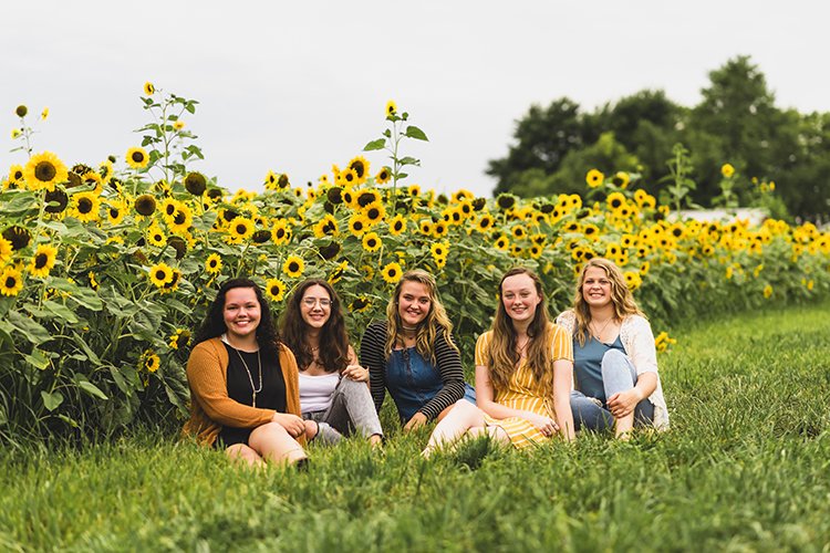 Senior representative team sitting on grass in front of a sunflower field, laughing together during a summer senior session in Connersville, Indiana.