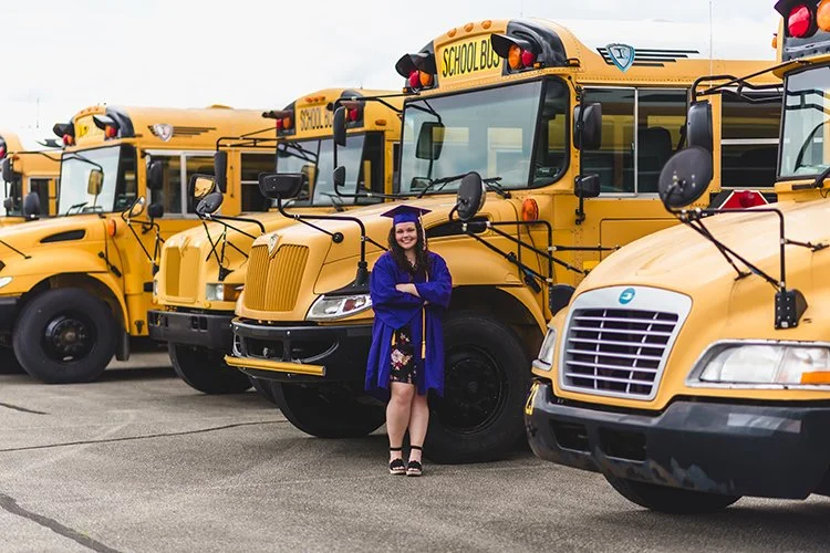 Graduating high school senior wearing a cap and gown, smiling with arms crossed in front of yellow school buses in Muncie, Indiana.